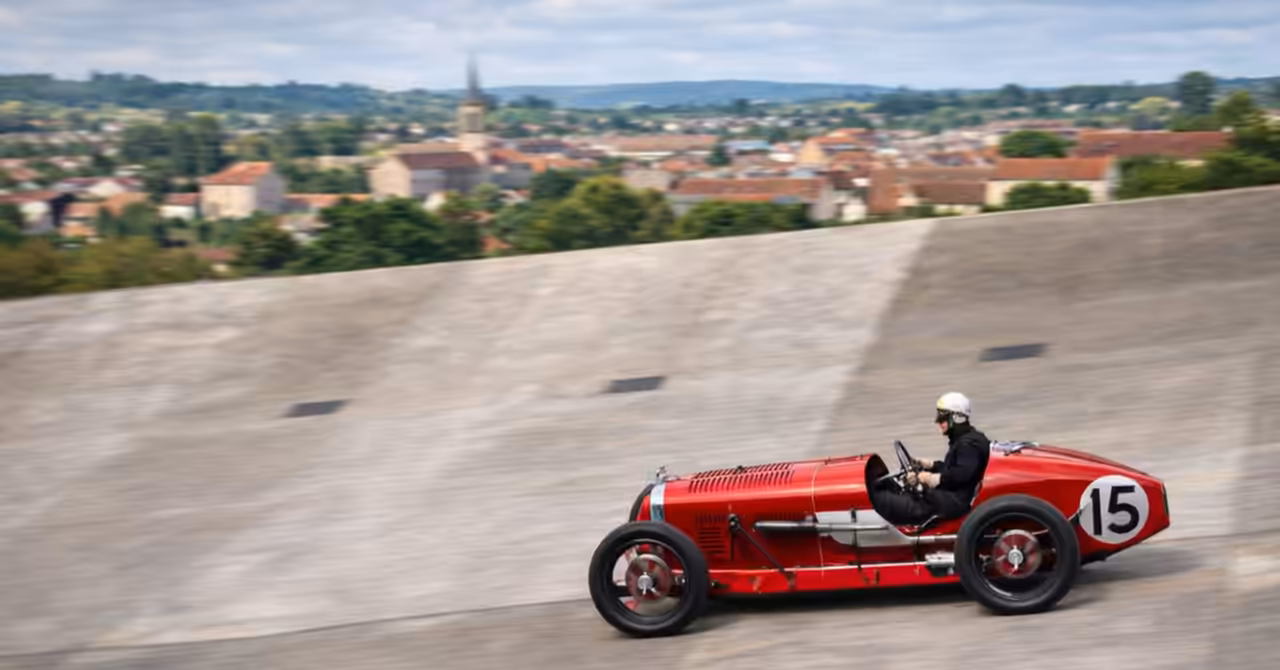 Artist’s impression of a red 1927 Amilcar Course 6 racing left to right on a steep concrete banking, with a French provincial town in the background.