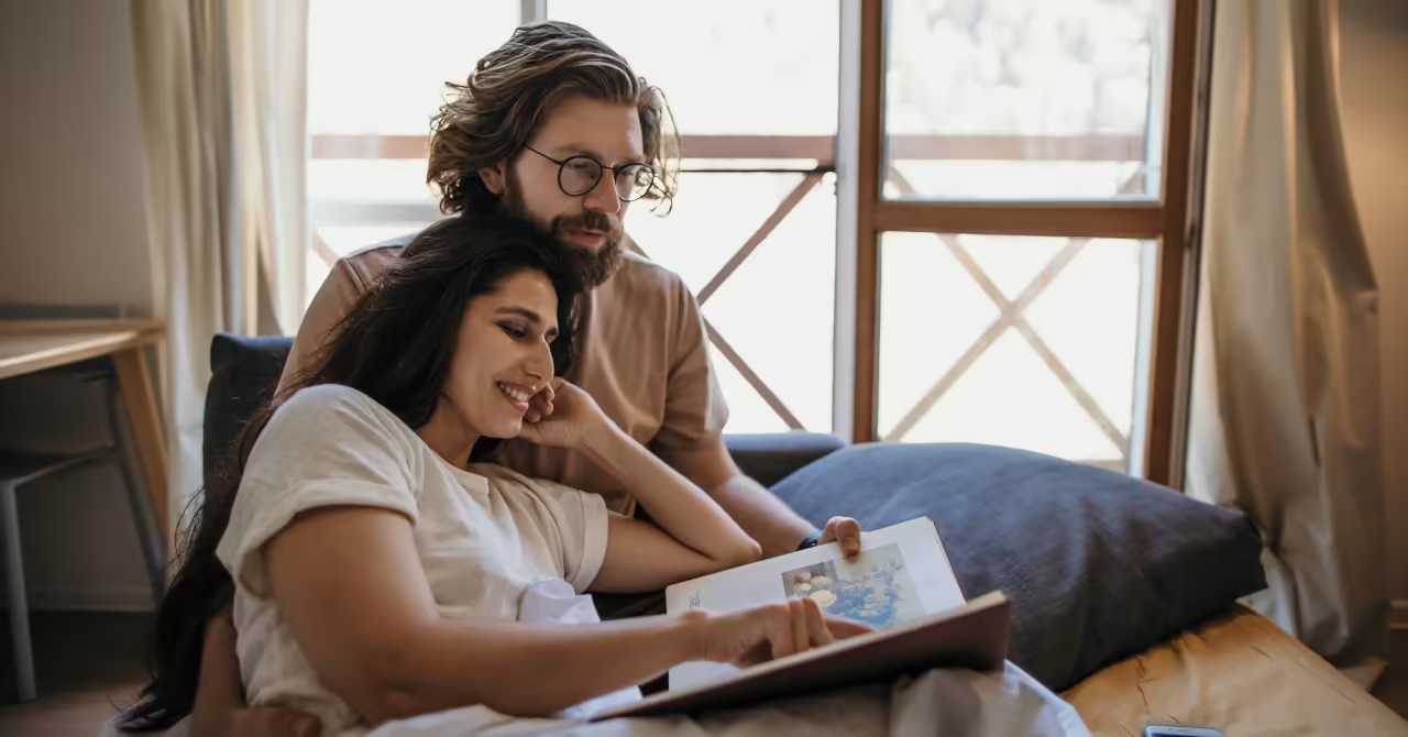 Two adults sitting closely on a sofa indoors, reading an illustrated book together near a window.